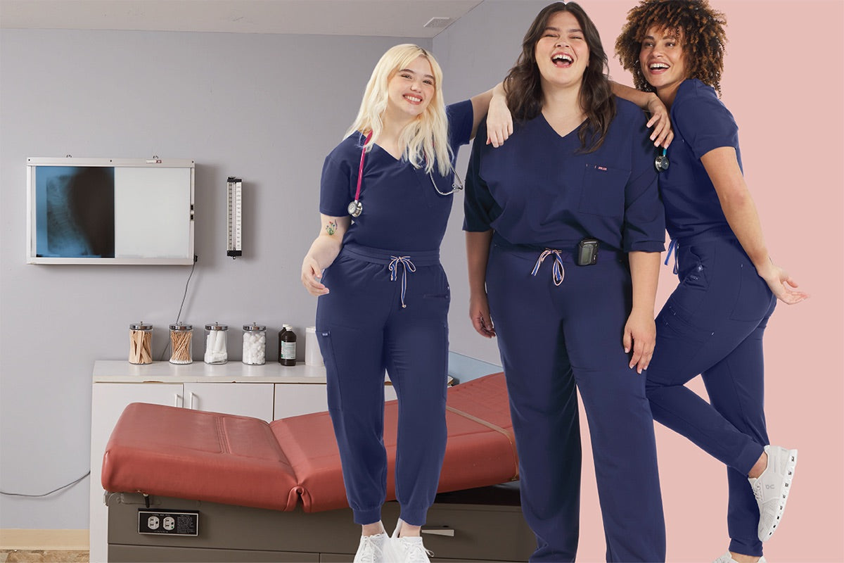 Three medical professionals in a clinic exam room wear navy scrub uniforms that look polished yet flexible, representing versatile scrubs designed for NPs and PAs who split time between clinic and hospital work.